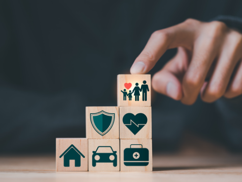 Wooden blocks with home, car, health and family icons stacked like steps while a hand places the top block